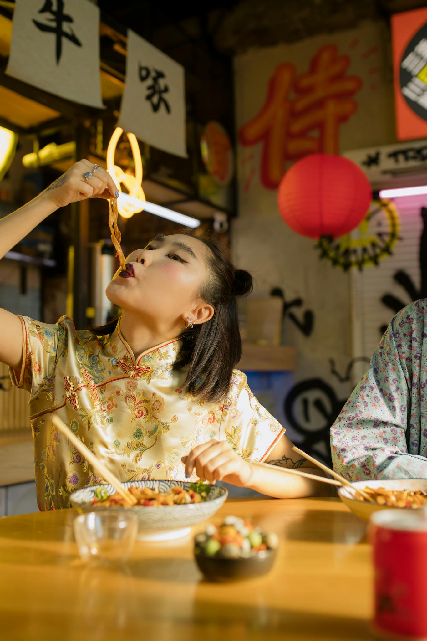 Woman in traditional dress savoring noodles with chopsticks in a vibrant Asian restaurant.